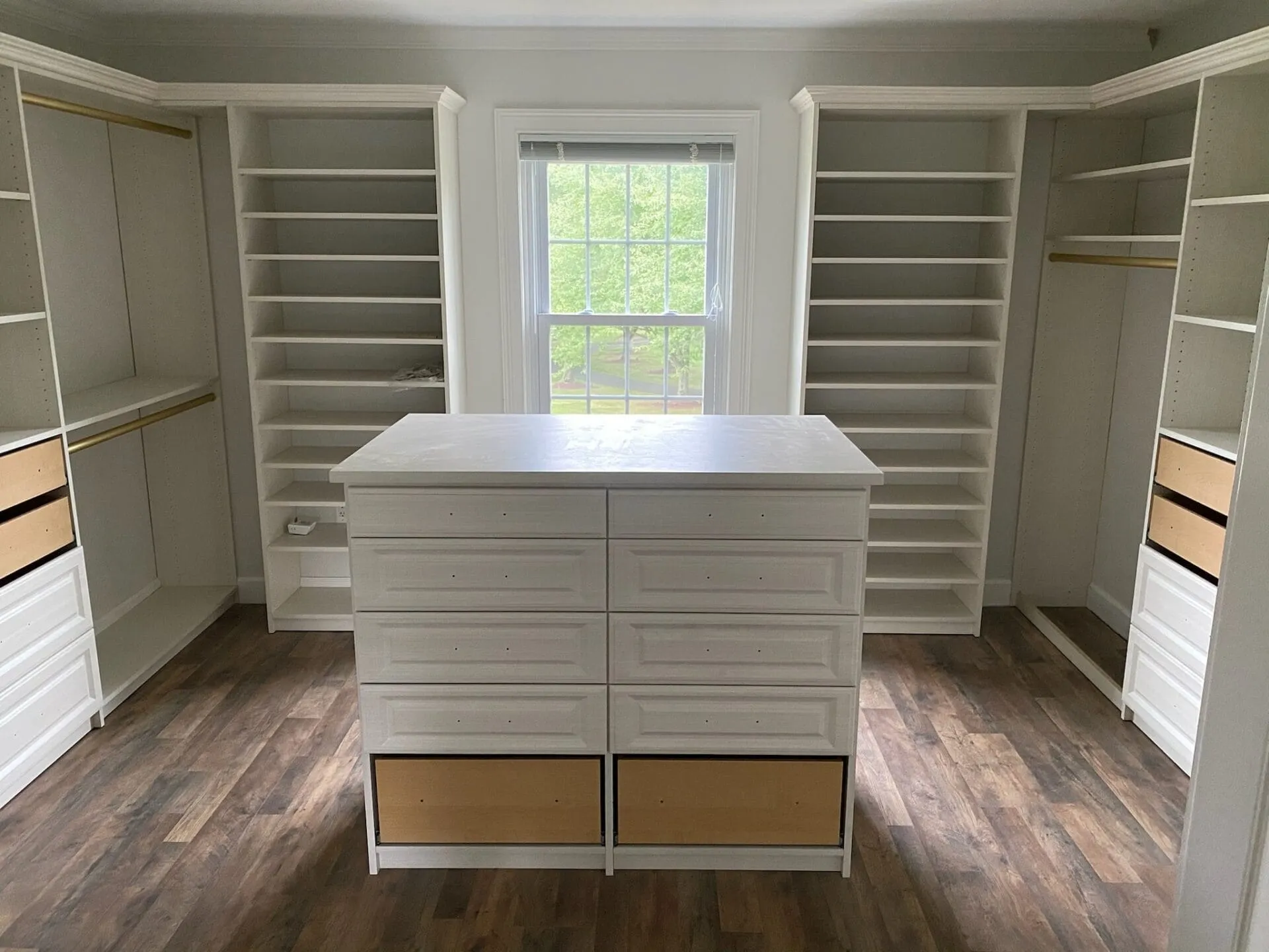 Spacious walk-in closet with white built-in shelving, hanging rods, and a central island dresser beneath a window, featuring wood-look flooring.