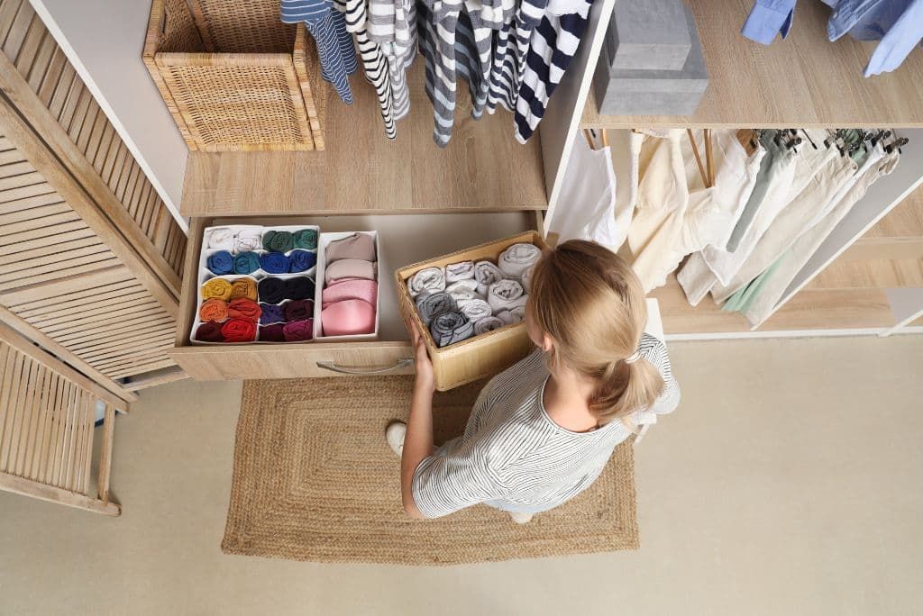 Overhead view of a woman organizing rolled clothing in a walk-in closet drawer, with neatly arranged garments and storage boxes.