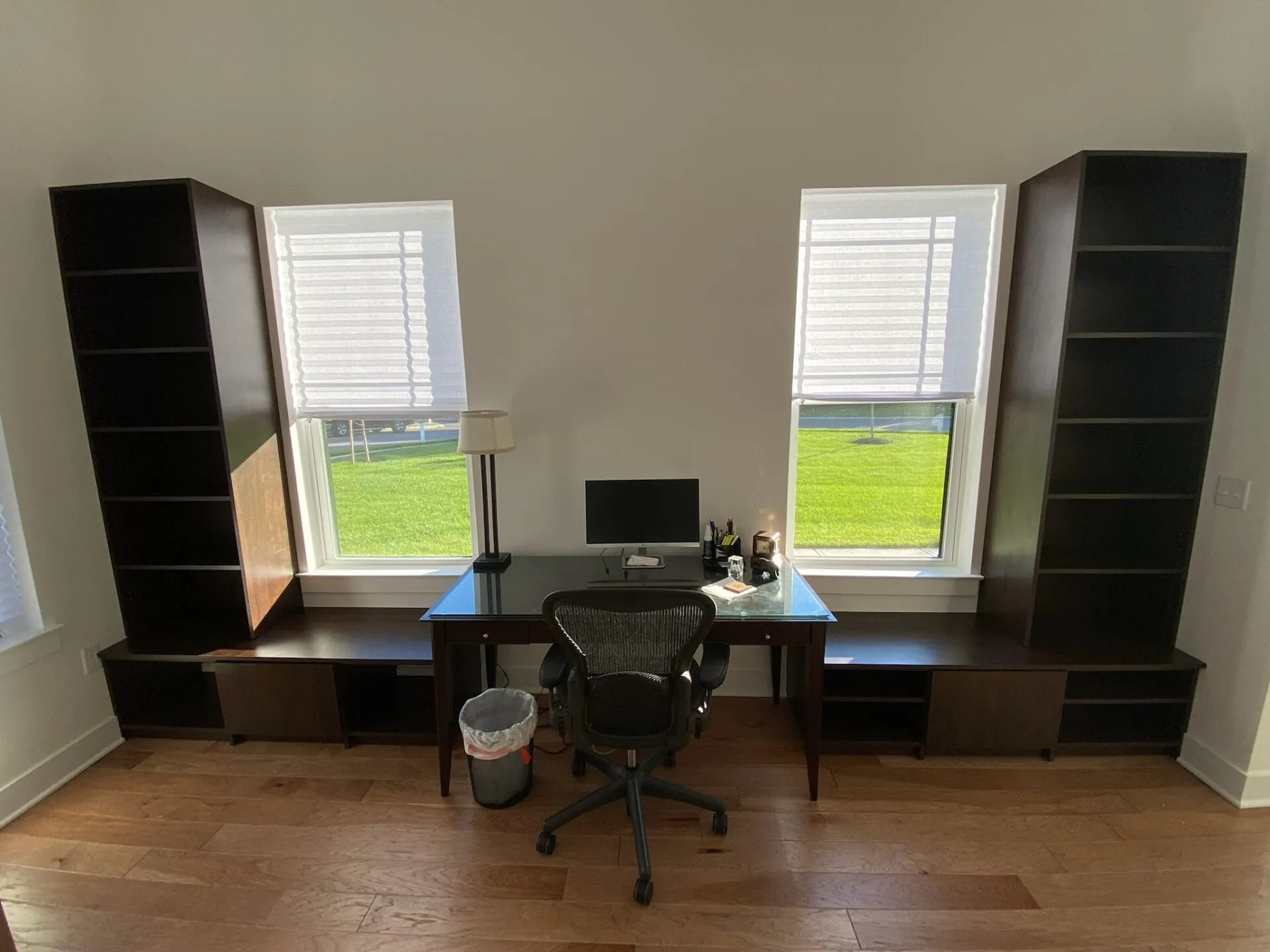 Modern home office setup with a desk centered between two windows, flanked by tall dark wood shelving units and hardwood floors.