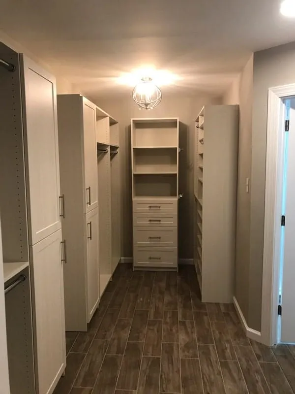 Walk-in closet with built-in white shelving, drawers, and hanging rods, featuring wood-look tile flooring and modern ceiling light.