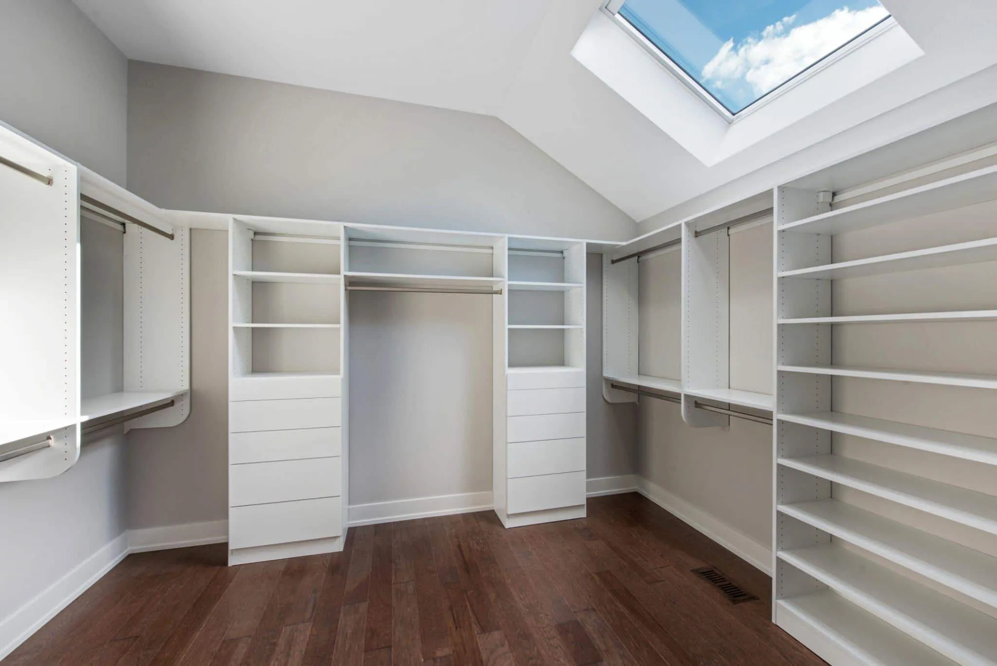 Spacious walk-in closet with white built-in shelving, drawers, and hanging rods along three walls, featuring a sloped ceiling and skylight above hardwood floors.