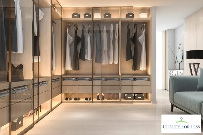 Modern walk-in closet with glass doors, wood shelving, hanging clothes, shoe storage, drawers, and warm built-in lighting for luxury wardrobe organization