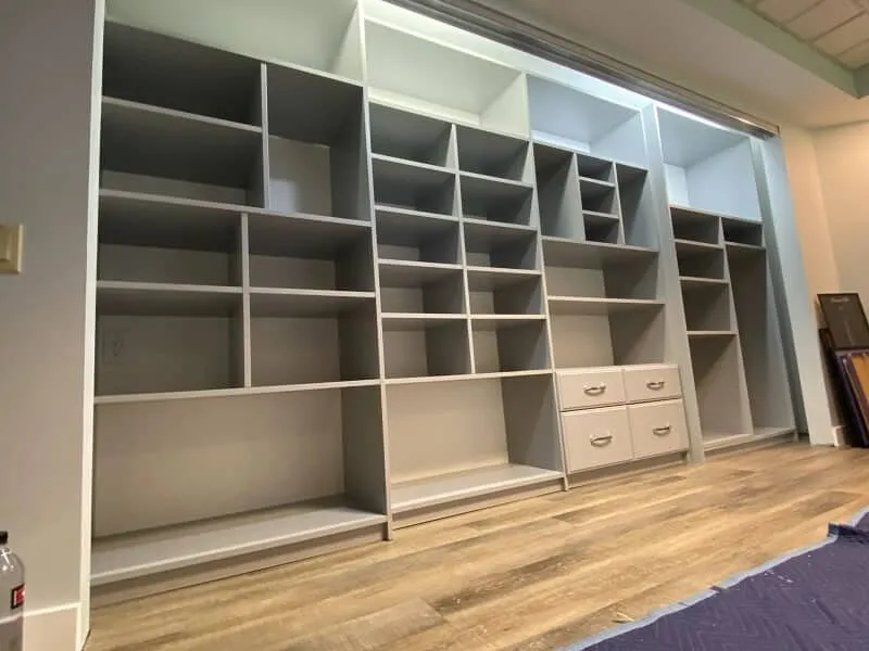Custom built-in closet organizer wall with open shelving cubbies, hanging sections, and drawers installed in a room with wood flooring.